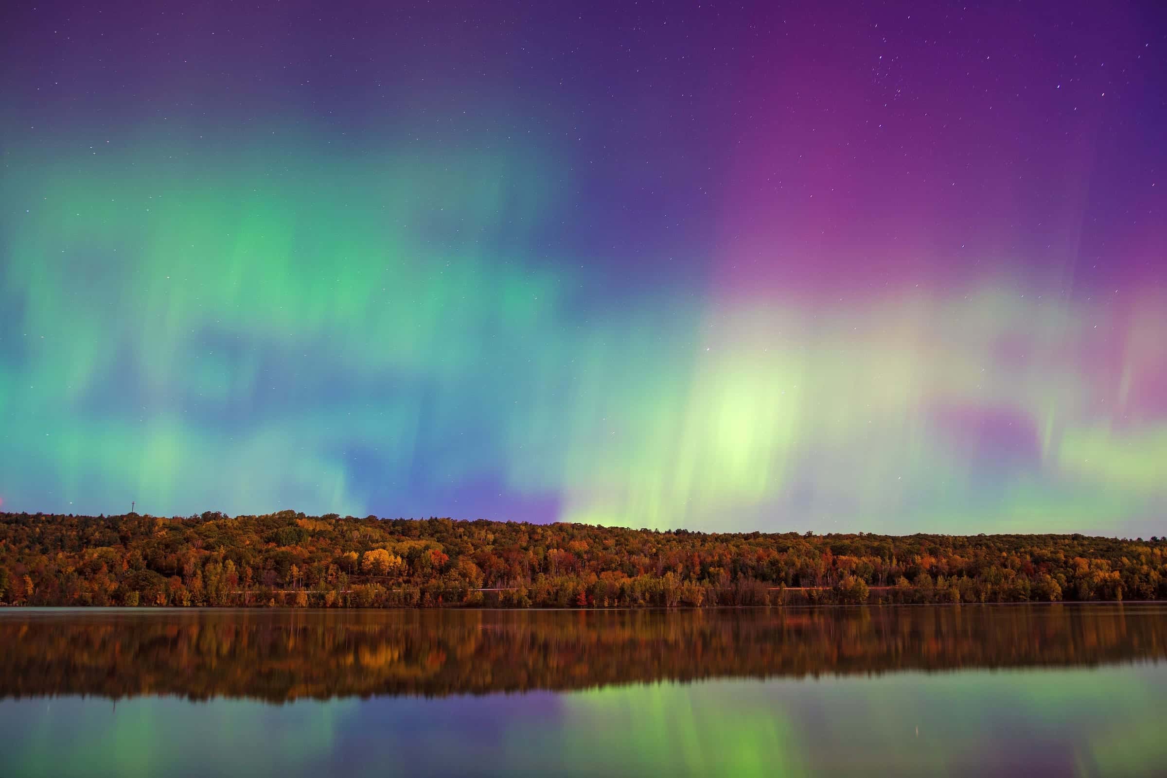 Green and magenta aurora over an autumn forest line, mirrored in a still lake