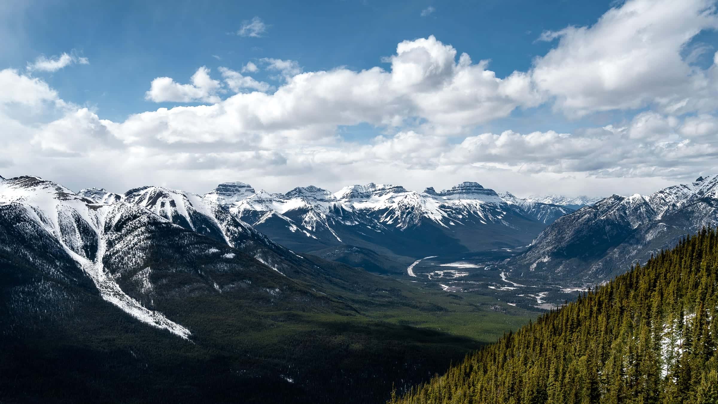 A wide alpine valley seen from a forested ridge, with snow-streaked peaks, a braided river and bright cumulus