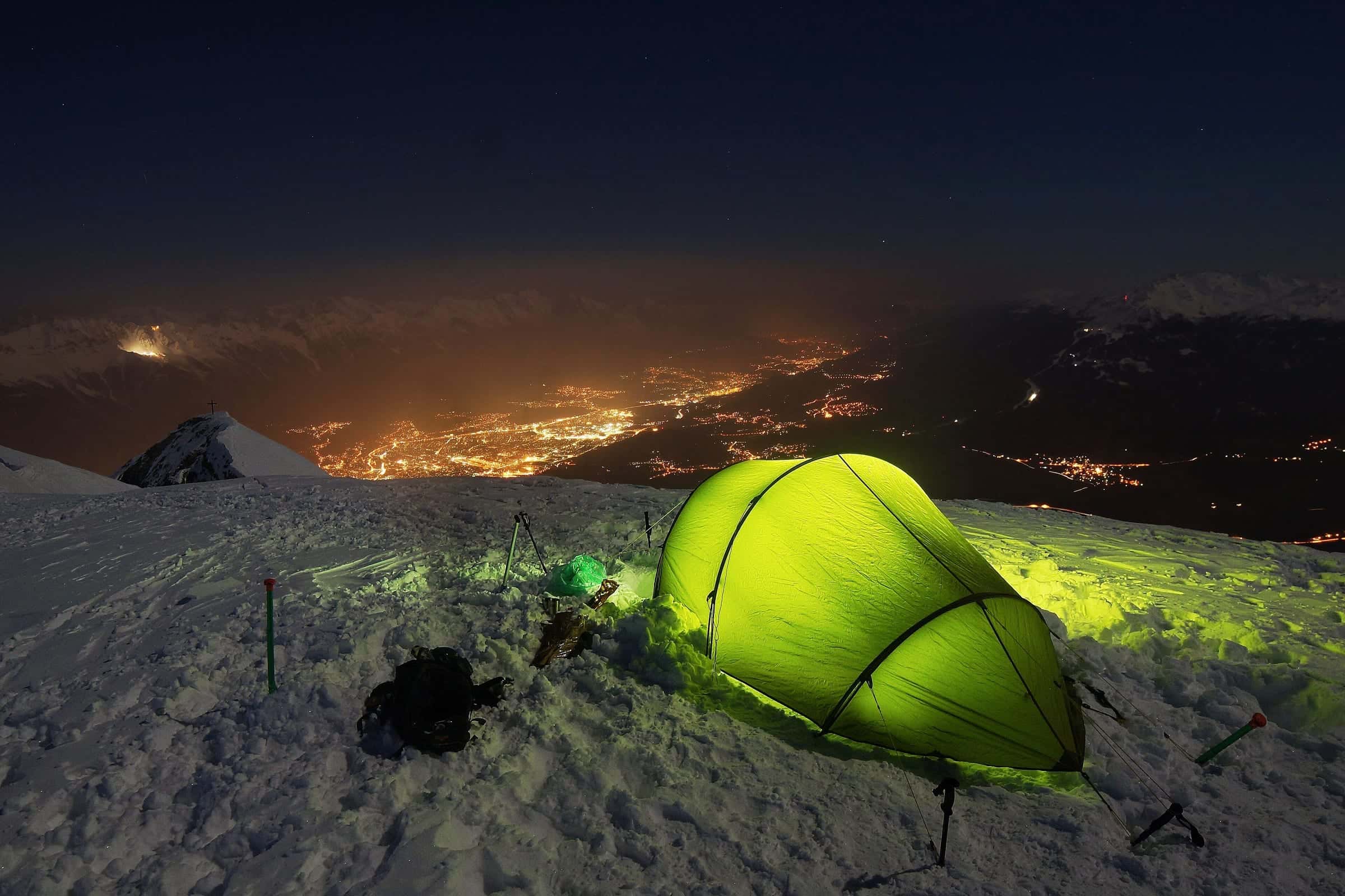 A glowing green tent on a snowy mountaintop at night, with lights in the valley far below