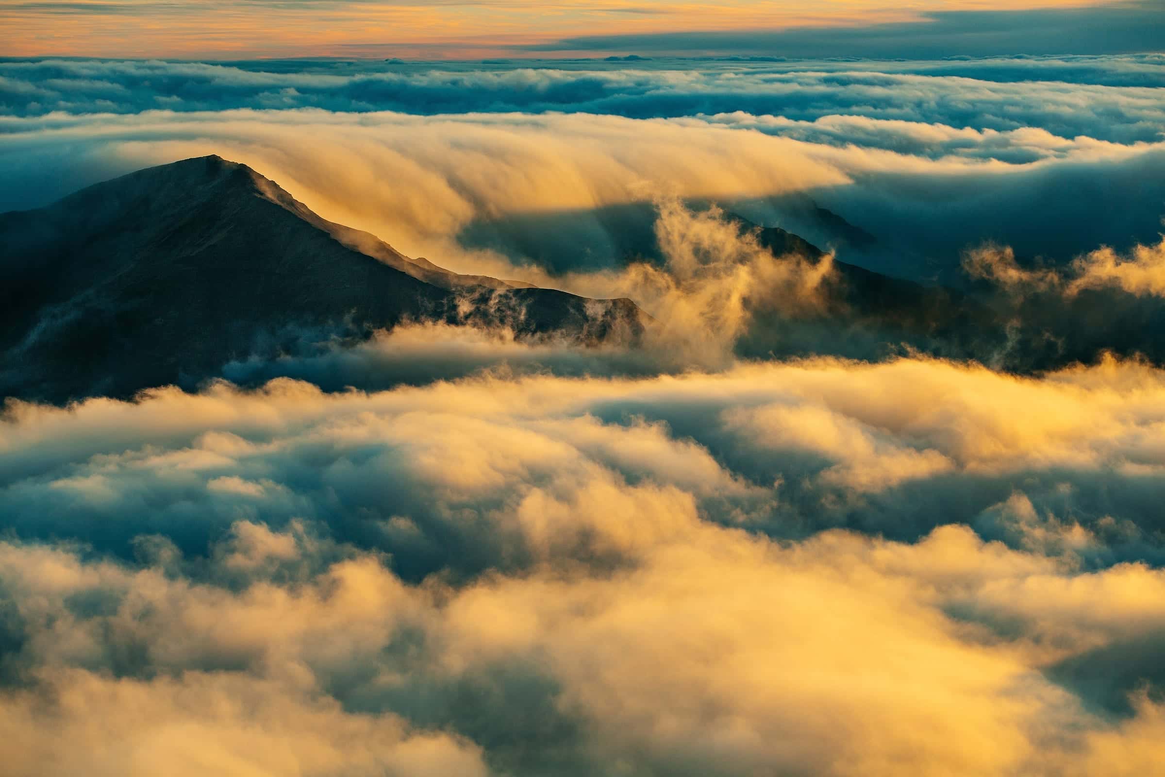 A dark ridge dropping into a golden, back-lit sea of cloud with teal shadows