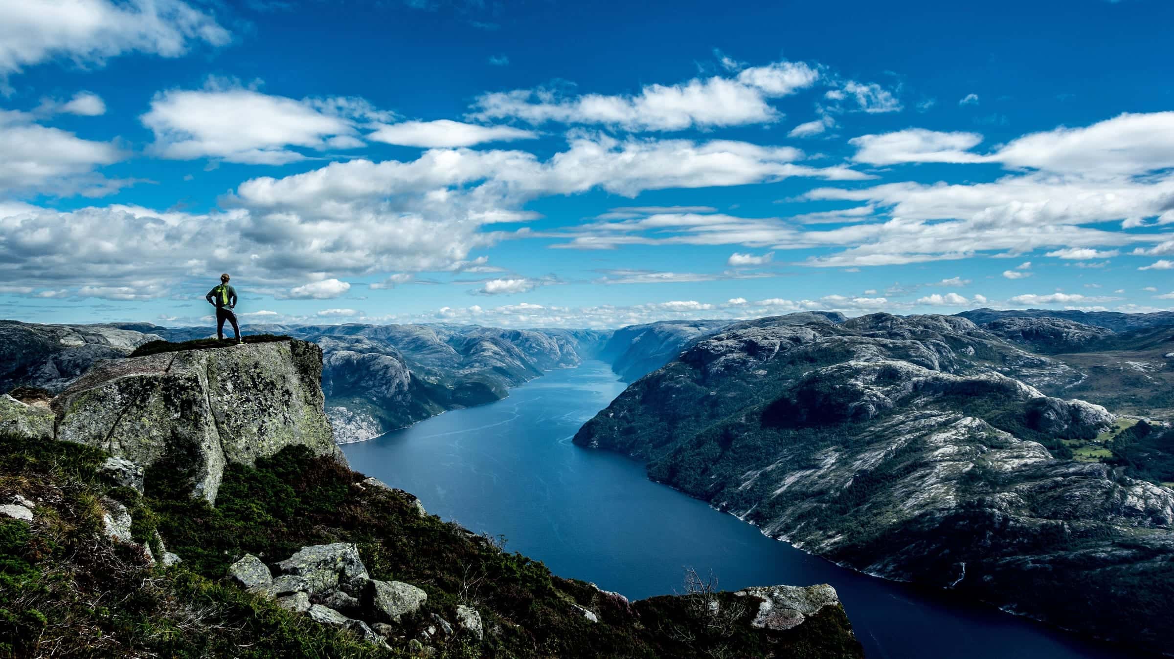 A small figure on a granite overlook above a deep-blue fjord under a sky full of cumulus